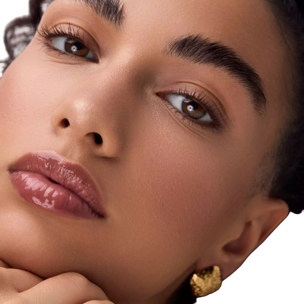 Nahaufnahme eines Gesichts mit Lippenöl. Die Lippen sind glänzend. Das Gesicht hat dunkle Augenbrauen und goldene Ohrringe.