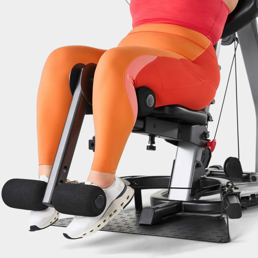 Beine einer Person auf einem Fitnessgerät. Schwarze Polster, orangefarbene Hose, weiße Schuhe.