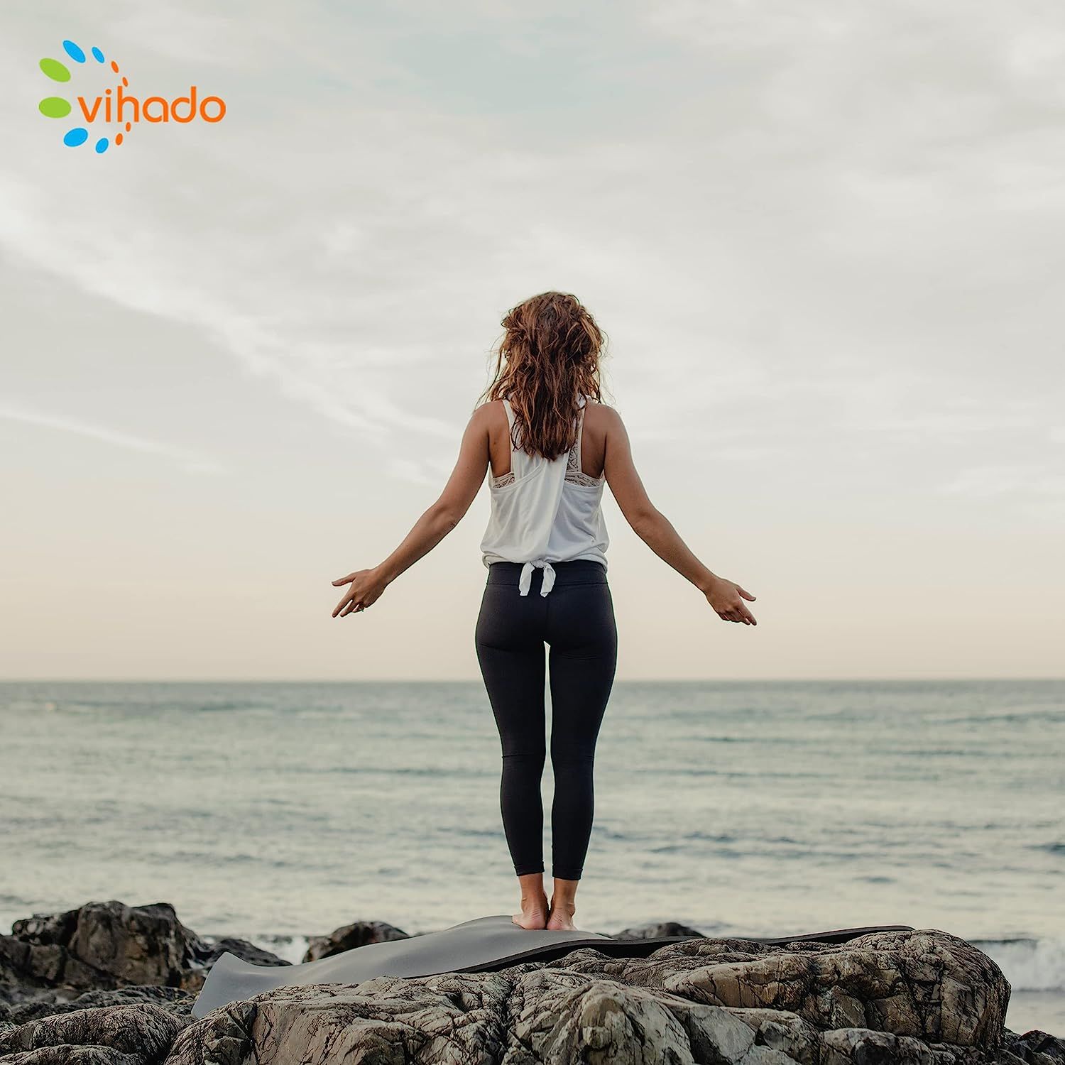 Eine Frau steht auf einem Felsen am Meer, mit ausgestreckten Armen. Sie trägt eine weiße Weste und schwarze Leggings. Der Hintergrund zeigt das Meer und den Himmel.
