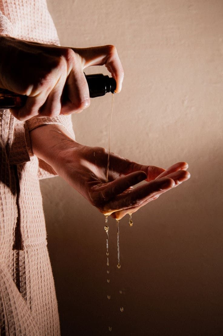 Hände halten eine schwarze Flasche. Öl wird auf die Handfläche gegossen. Hintergrund: beige.