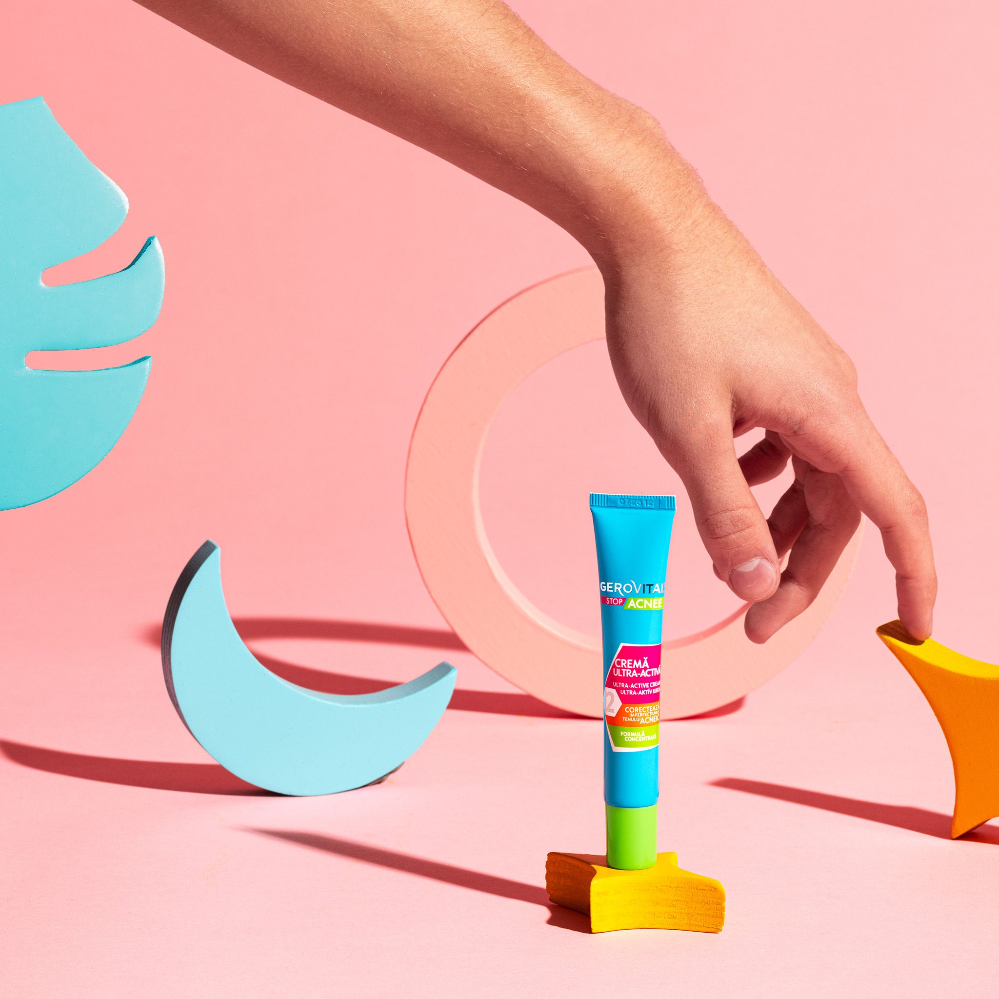 Türkis-grüne Tube auf gelbem Podest, gehalten von einer Hand. Rosa Hintergrund mit Holzformen. Aufschrift: Stop Acnee.