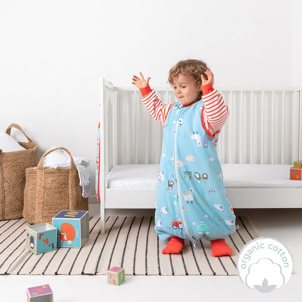 Kind trägt blauen Schlafsack mit Bauernhof-Motiv. Kind steht vor weißem Bett. Aufdruck: organic cotton.