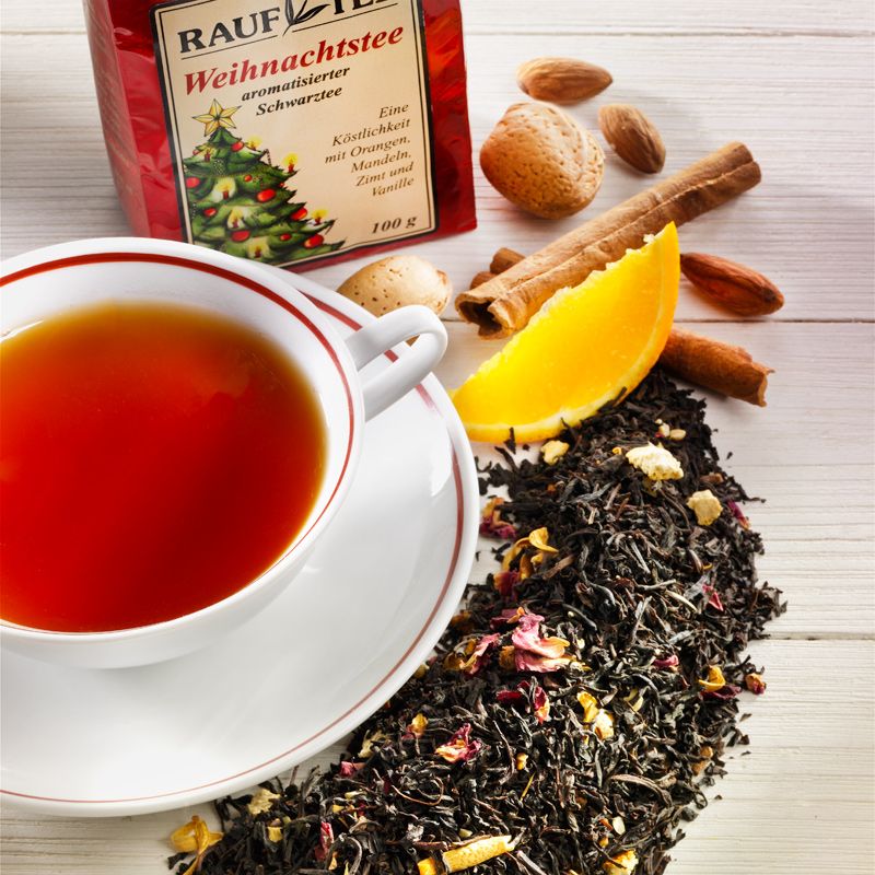 Tasse Tee mit Zutaten: Zimt, Mandeln, Orange. Rauf Tee Weihnachtstee-Packung im Hintergrund.