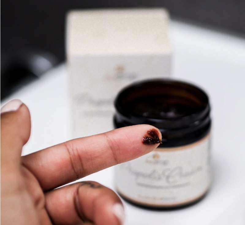 Finger mit Creme. Dose "Propolis Cream" im Hintergrund. Creme dunkelbraun. Dose mit "be drop" Logo. Enthält Bienenwachs und Traubenkernöl.
