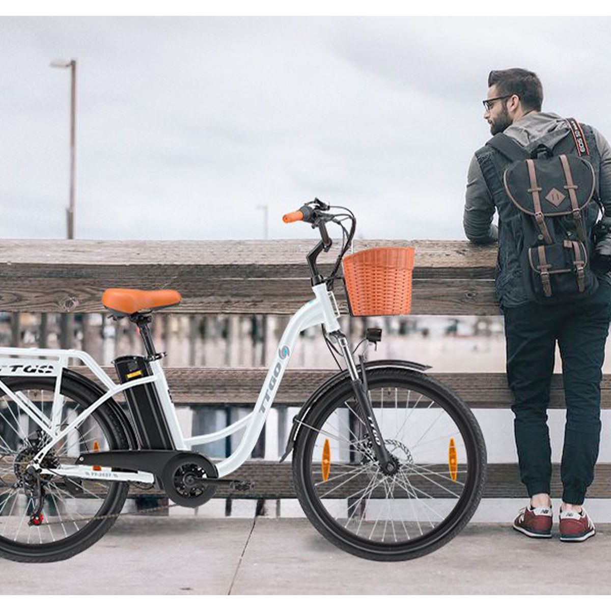 Person steht neben weißem Elektrofahrrad mit Korb. Brauner Sattel und Lenkergriffe. TTGO-Logo. Hintergrund: Steg.