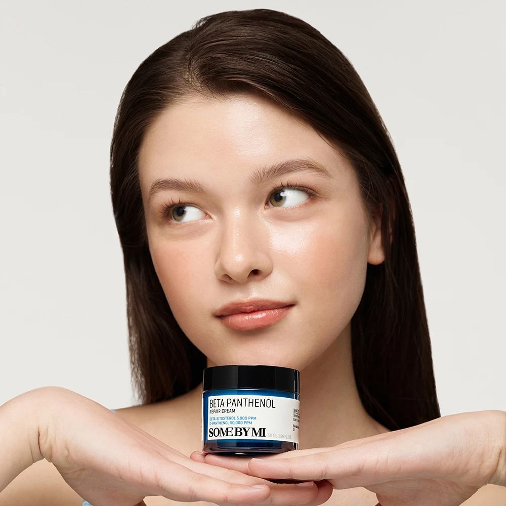 Eine Frau hält eine blaue Creme-Dose mit schwarzem Deckel. Auf der Dose steht "BETA PANTENOL" und "SOME BY MI".