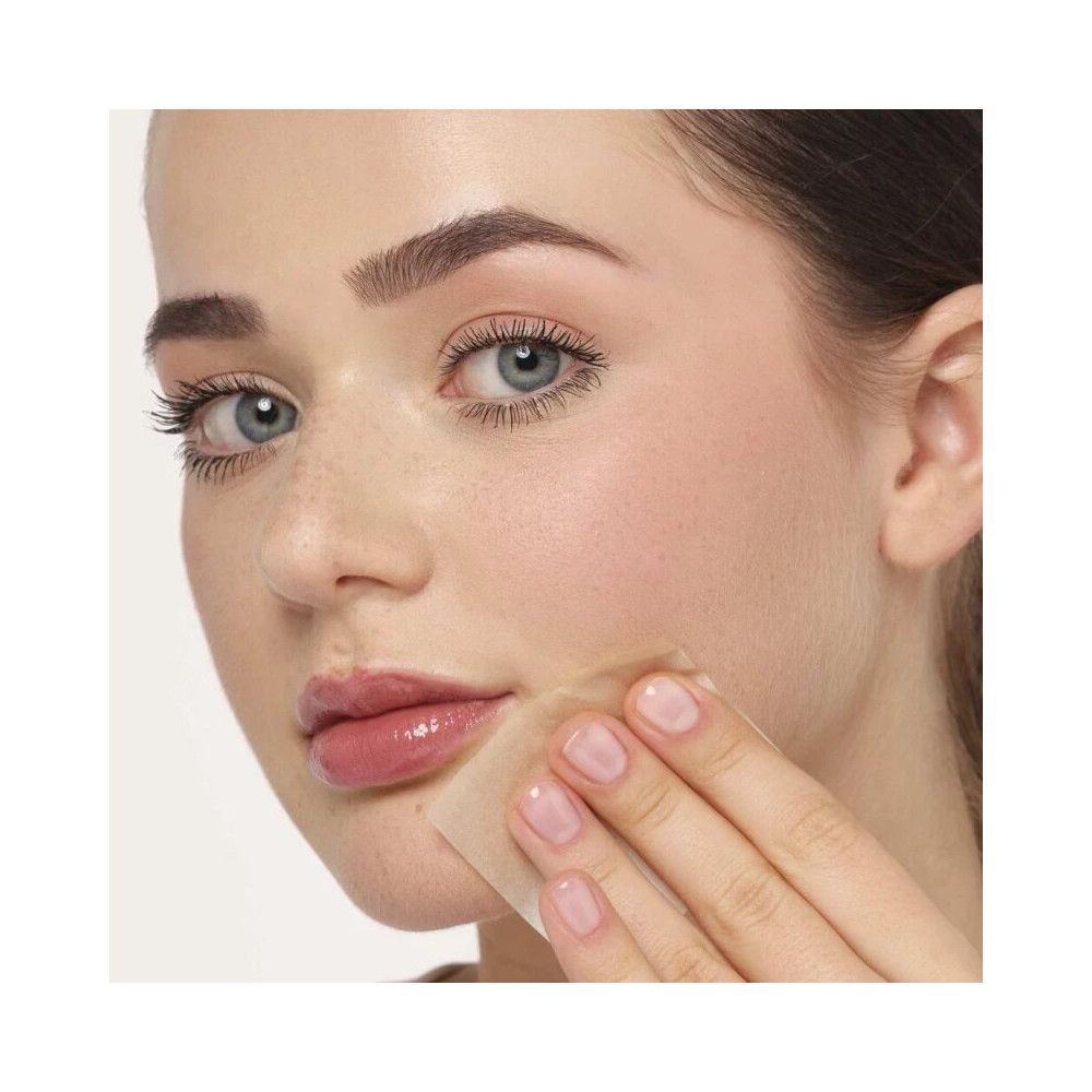 Frau mit hellem Teint, benutzt ein quadratisches Ölpapier auf der Haut. Das Papier berührt die Wange.