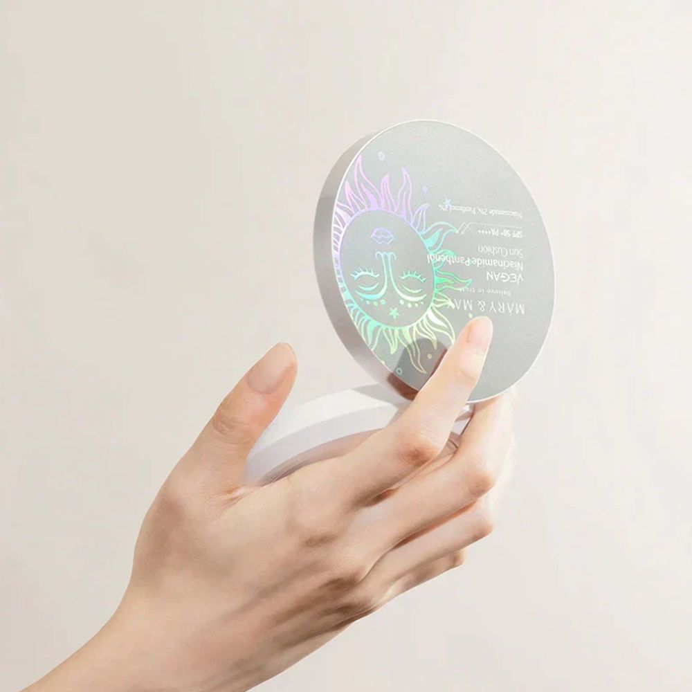 Runder, offener Behälter, gehalten von einer Hand. Holografische Sonne auf dem Deckel. Text: MARY & MAY, Vegan Niacinamide Panthenol Sun Cushion.