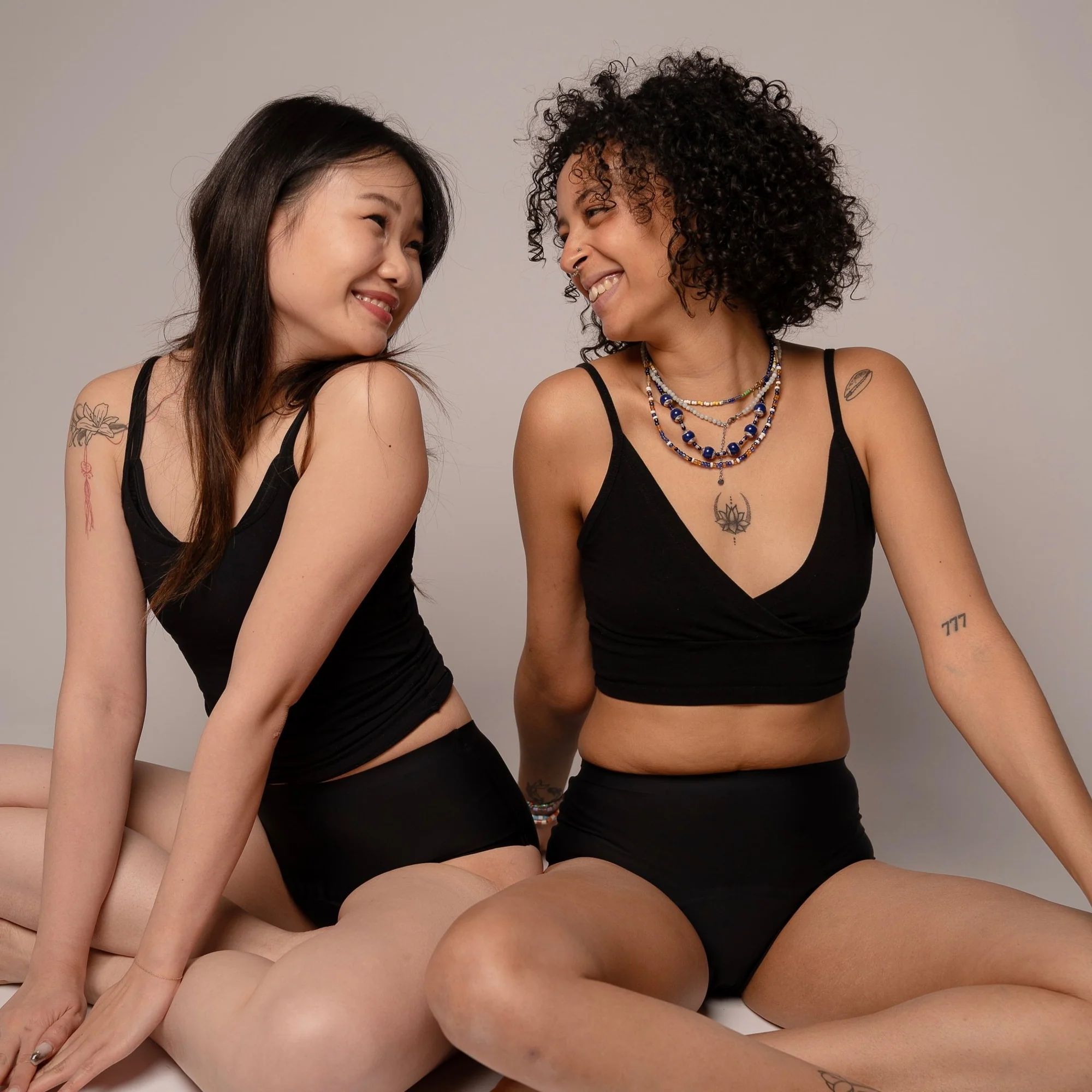 Zwei Frauen sitzen nebeneinander. Beide tragen schwarze Tops und Slips. Sie lächeln.