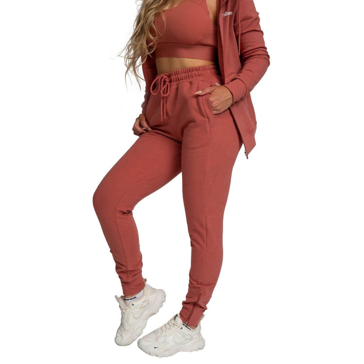 Damen Jogginghose in Zimtfarbe, schmal geschnitten, mit Kordelzug und Taschen, kombiniert mit Sport-BH und Hoodie.