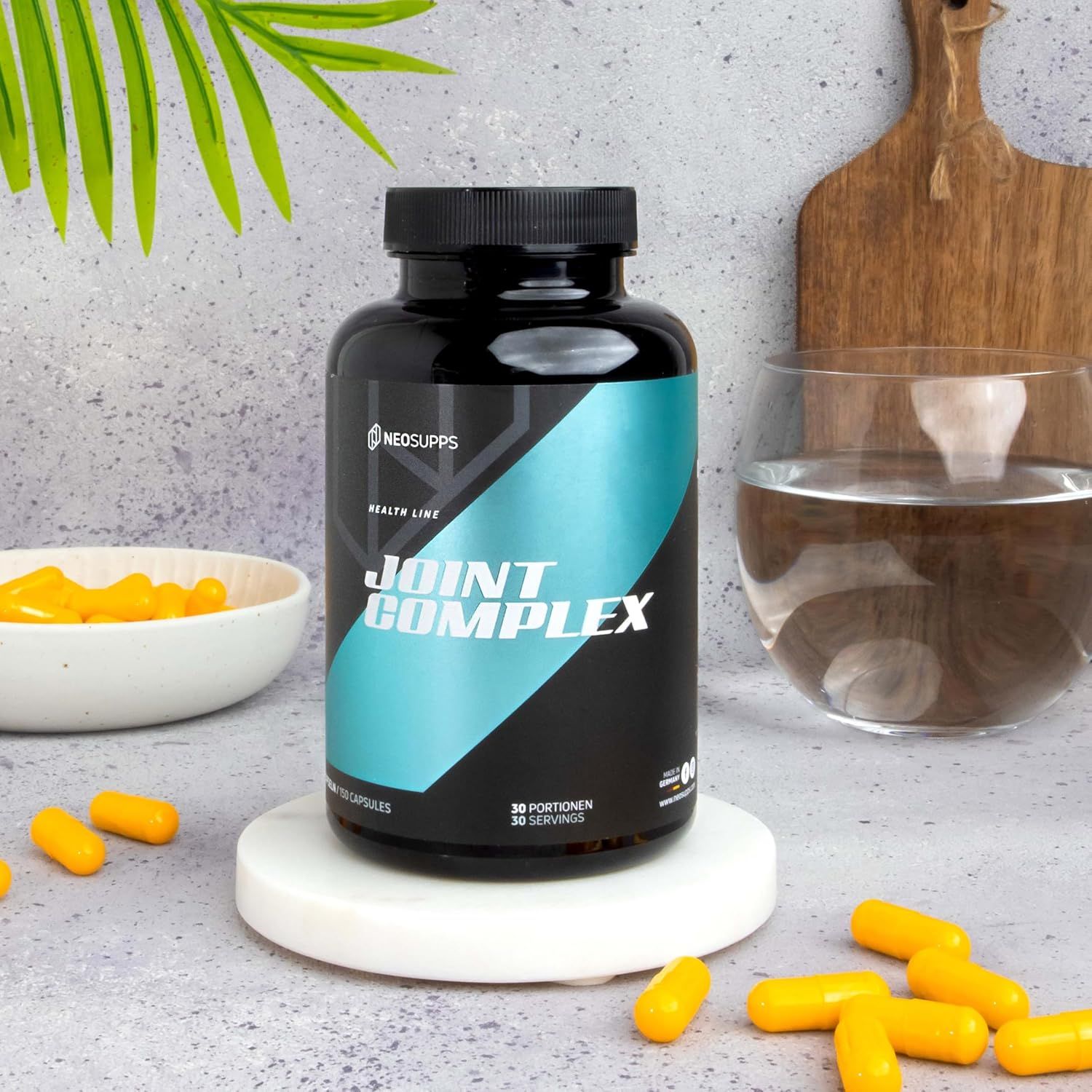 Schwarze Flasche "JOINT COMPLEX" auf weißem Podest. Gelbe Kapseln daneben. Glas Wasser, grüne Blätter im Hintergrund.
