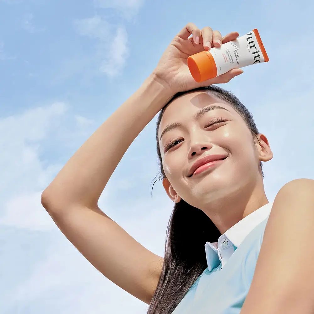 Frau hält eine Tube Sonnencreme über ihrem Kopf. Blauer Himmel im Hintergrund. Produkt: PURITO.