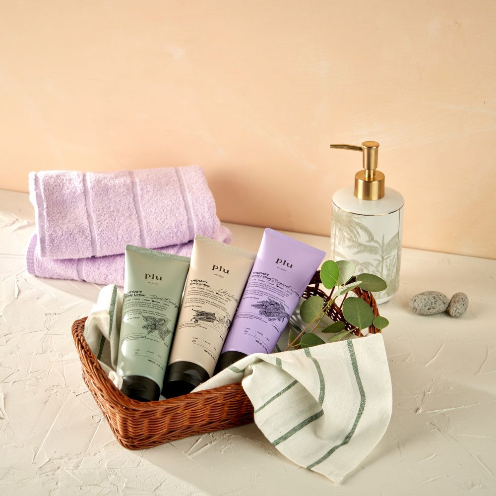 Korb mit drei Tuben 'plu AROMA Body Lotion' in verschiedenen Farben, Handtüchern und Seifenspender.