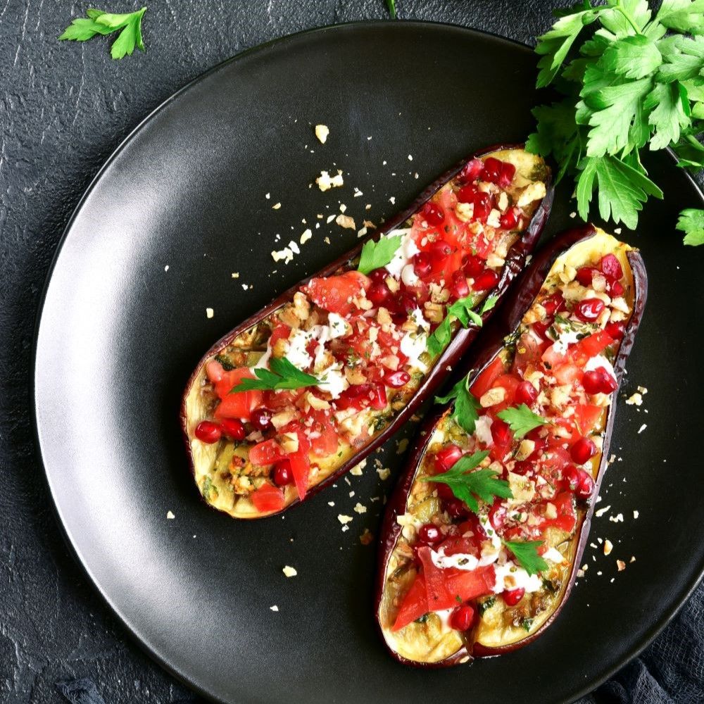 Zwei gefüllte Auberginen-Hälften auf schwarzem Teller. Belag mit Tomaten, Granatapfelkernen und Kräutern. Draufsicht.