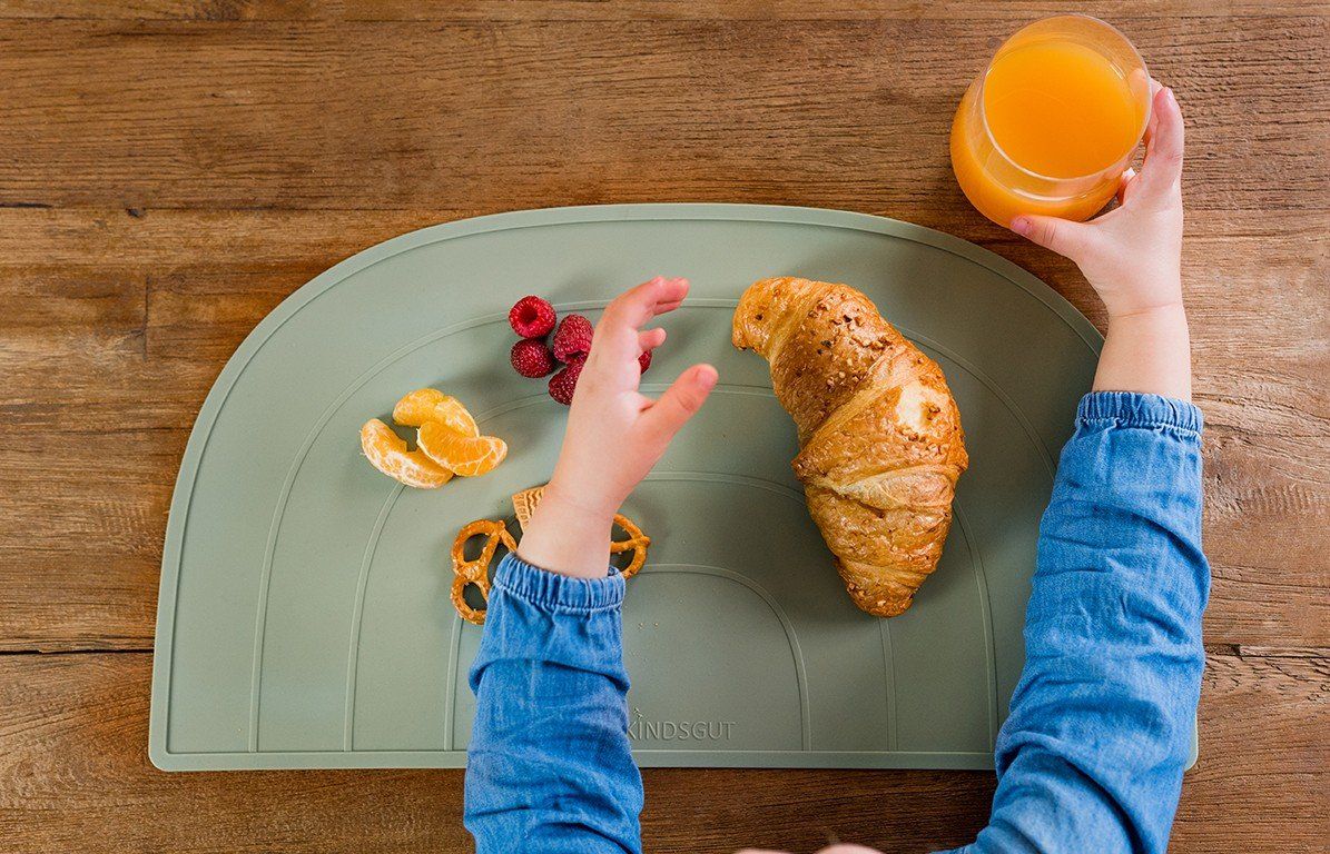 Kind greift nach Essen auf dem Platzdeckchen. Croissant, Obst und Brezeln sind sichtbar.
