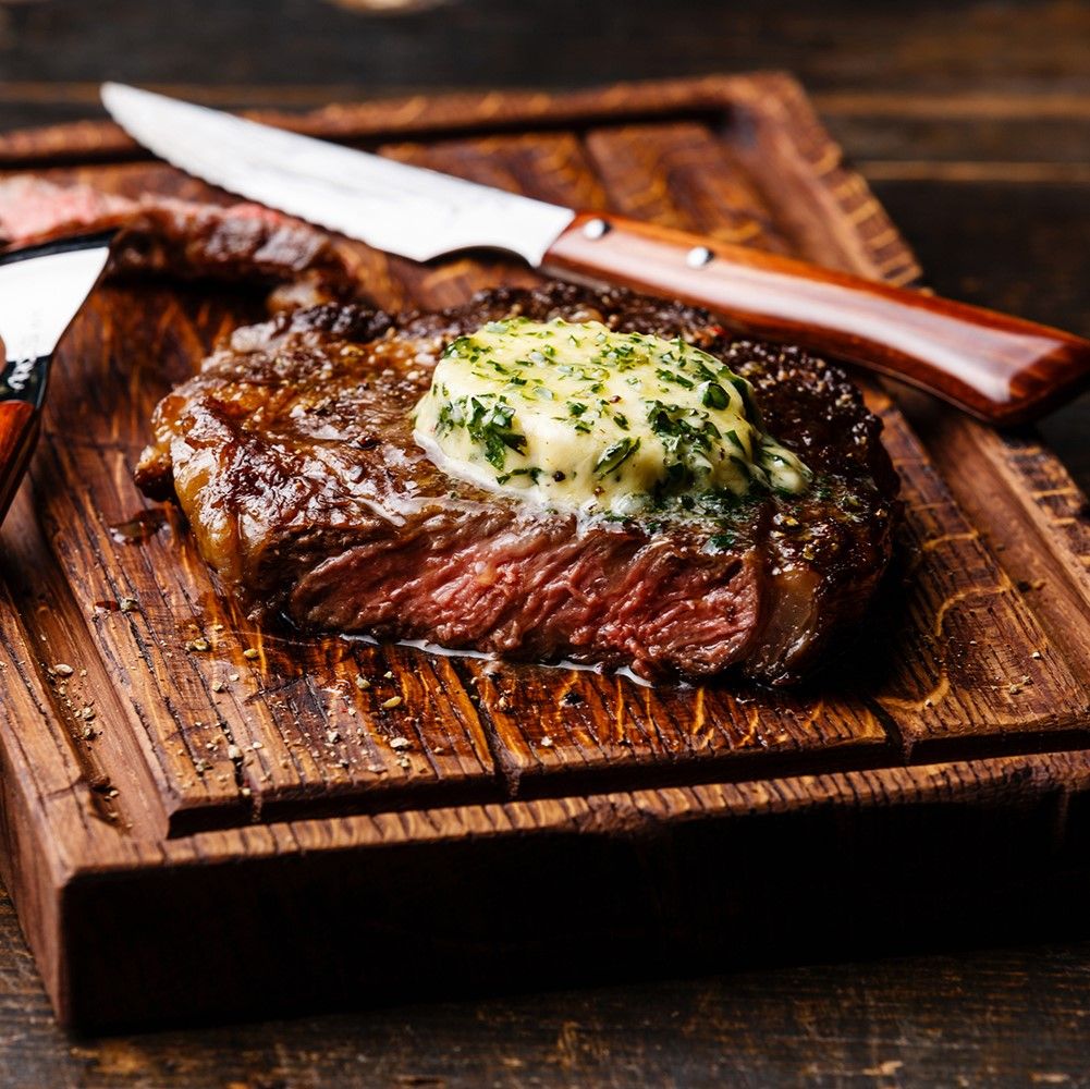 Steak auf Holzbrett mit Kräuterbutter. Steakmesser liegt daneben. Fleisch rot, Butter grün.