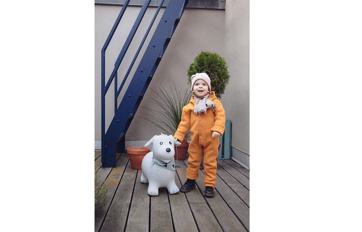 Kind steht neben einem grauen Hüpfhund. Das Kind trägt einen gelben Overall und eine Mütze. Der Hund hat ein Halstuch.
