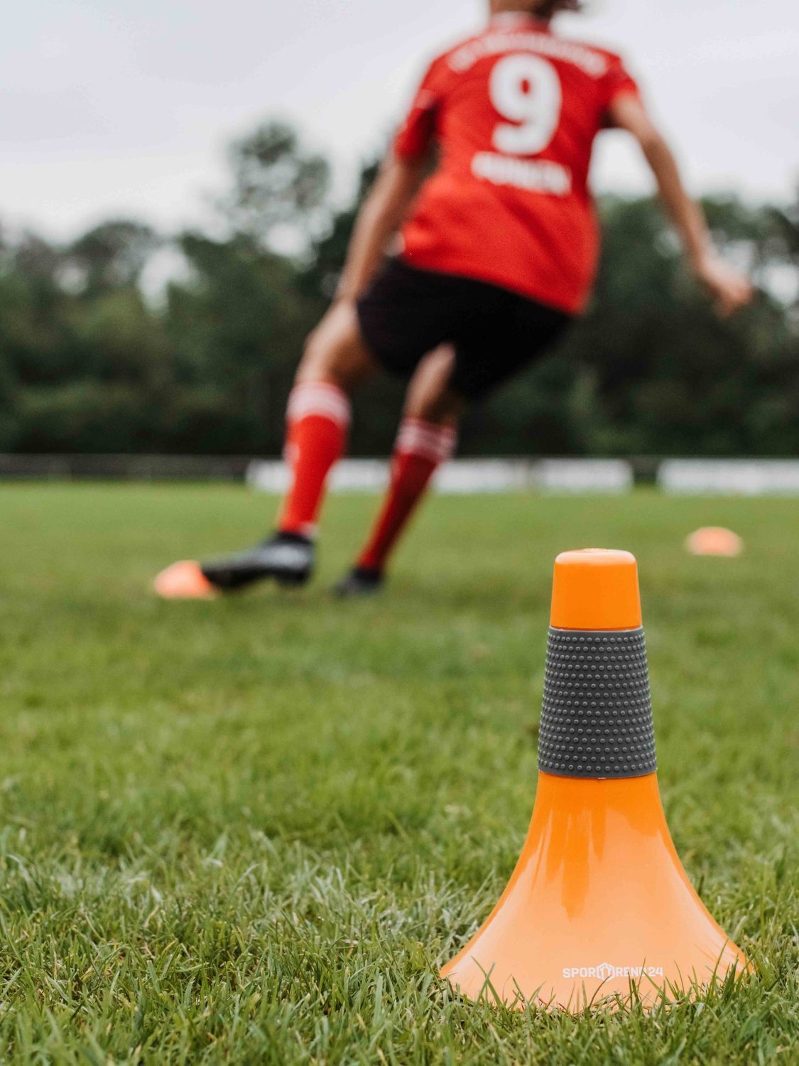 Orangener Speed Cone auf Rasen. Im Hintergrund ein Fußballspieler.