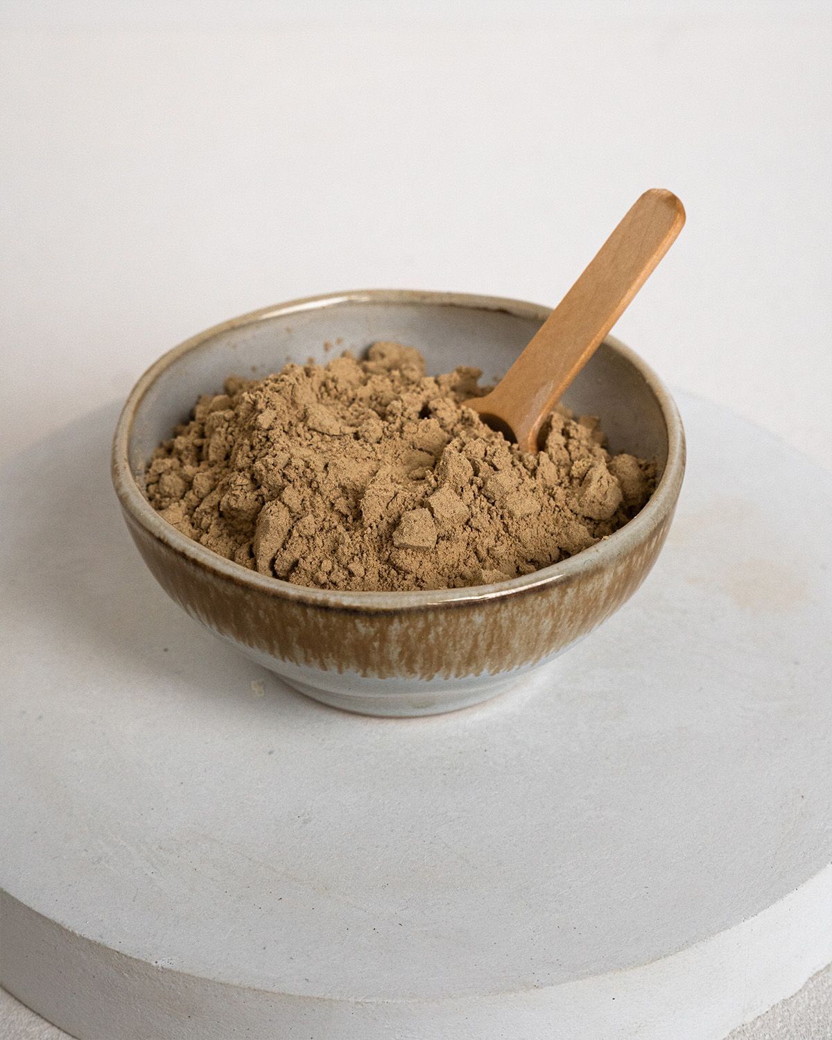 Braune Pulver in einer kleinen, hellbraunen Schale mit einem Holz-Löffel. Auf einer weißen Oberfläche.