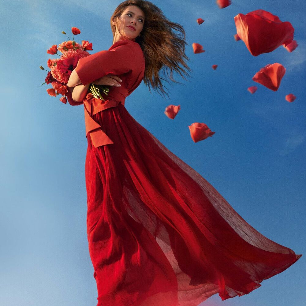 Frau in rotem Kleid mit Blumenstrauß. Rote Blütenblätter fliegen im blauen Himmel.