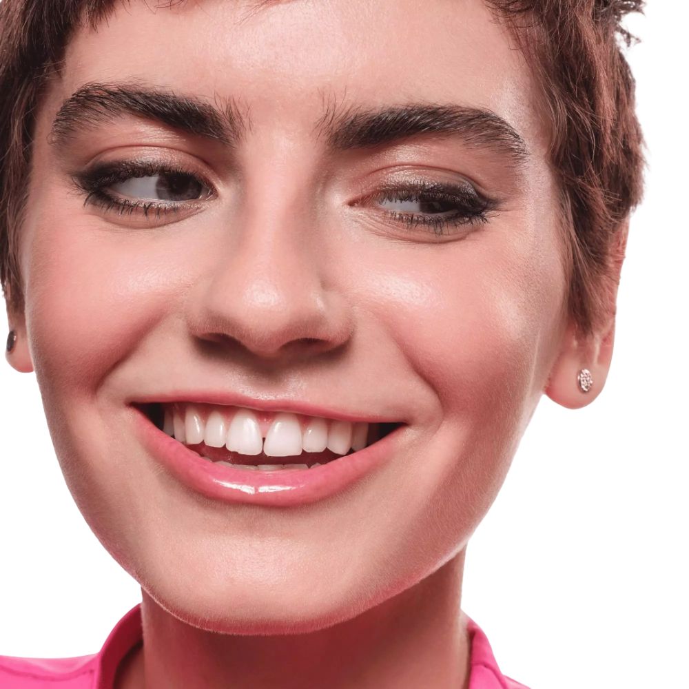 Porträt einer lächelnden Frau. Sie trägt schwarzen Eyeliner. Rosa Oberteil. Weißer Hintergrund.