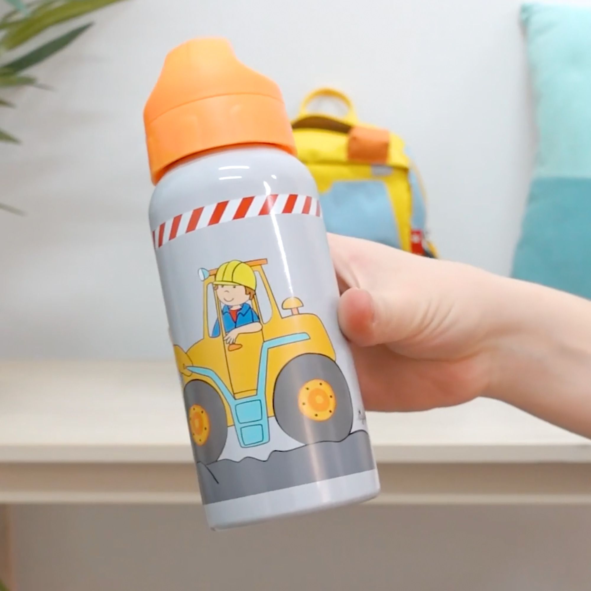 Trinkflasche mit orangefarbenem Deckel. Graue Flasche mit Bauarbeiter-Motiv. Gehalten von einer Hand.