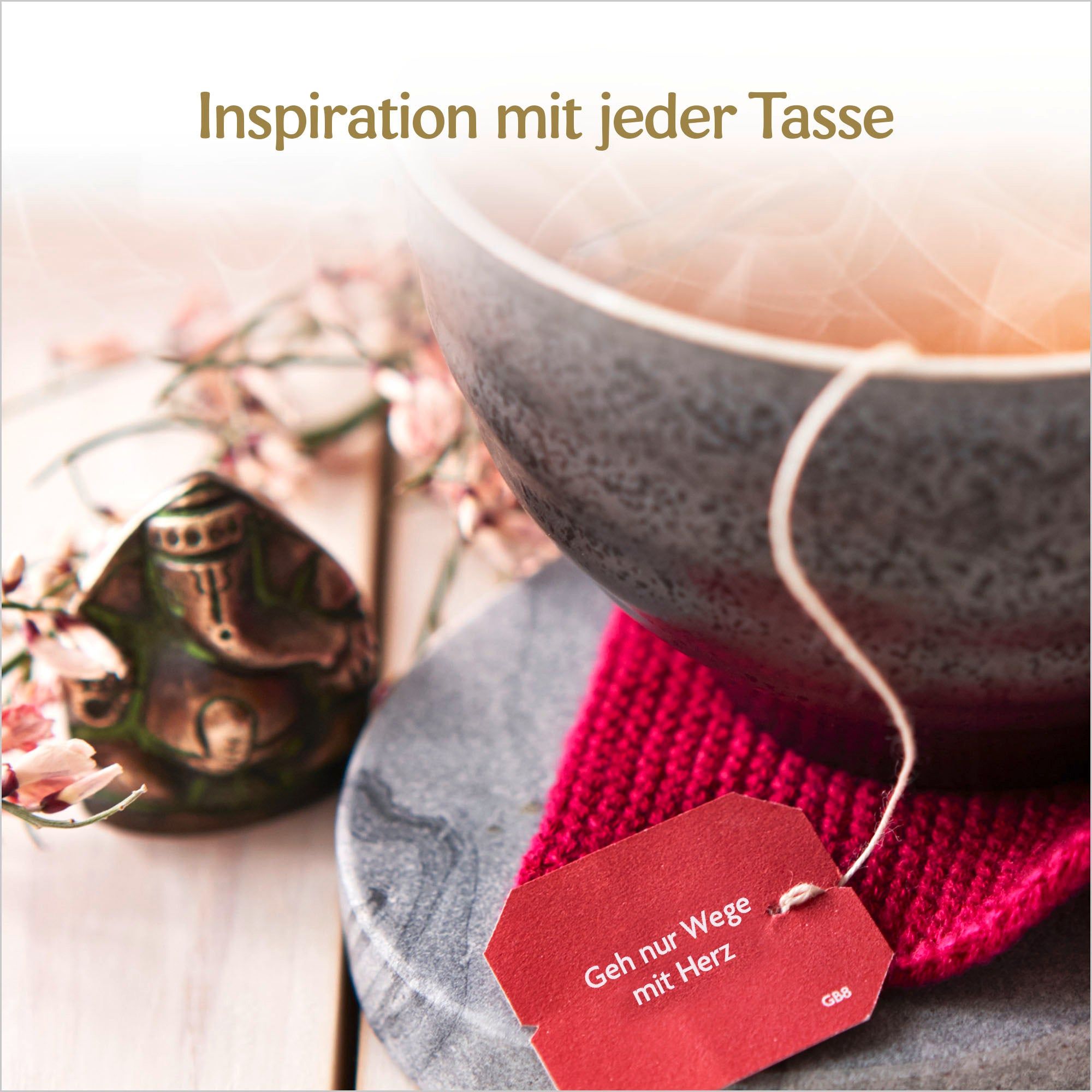 Tasse Tee mit Teebeutel. Roter Anhänger mit Text. Dekoration: kleine Statue, rosa Blüten. Text: Inspiration mit jeder Tasse.
