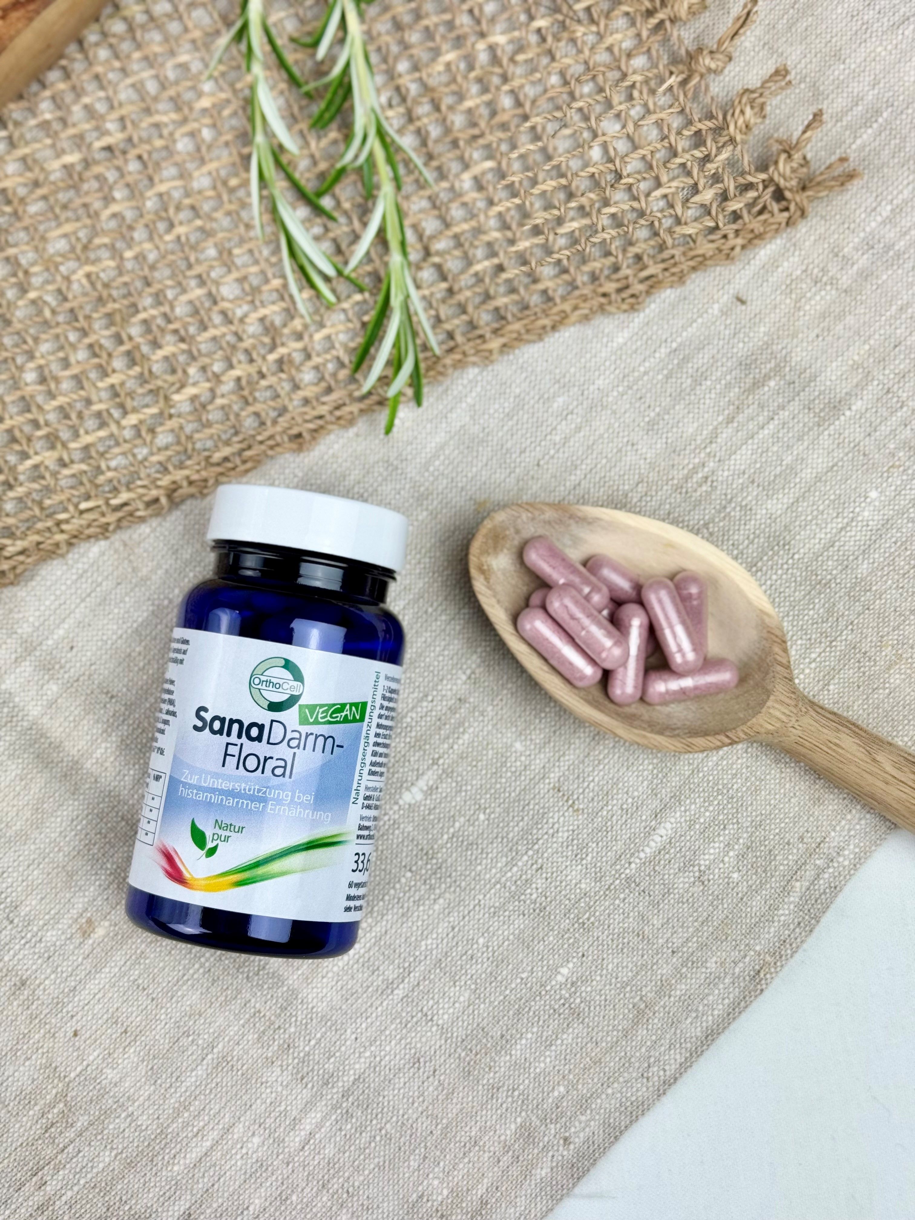 Blaue Flasche SanaDarm-Floral neben Holzlöffel mit Kapseln. Auf Leinenstoff. Vegan-Siegel. Rosmarinzweig.