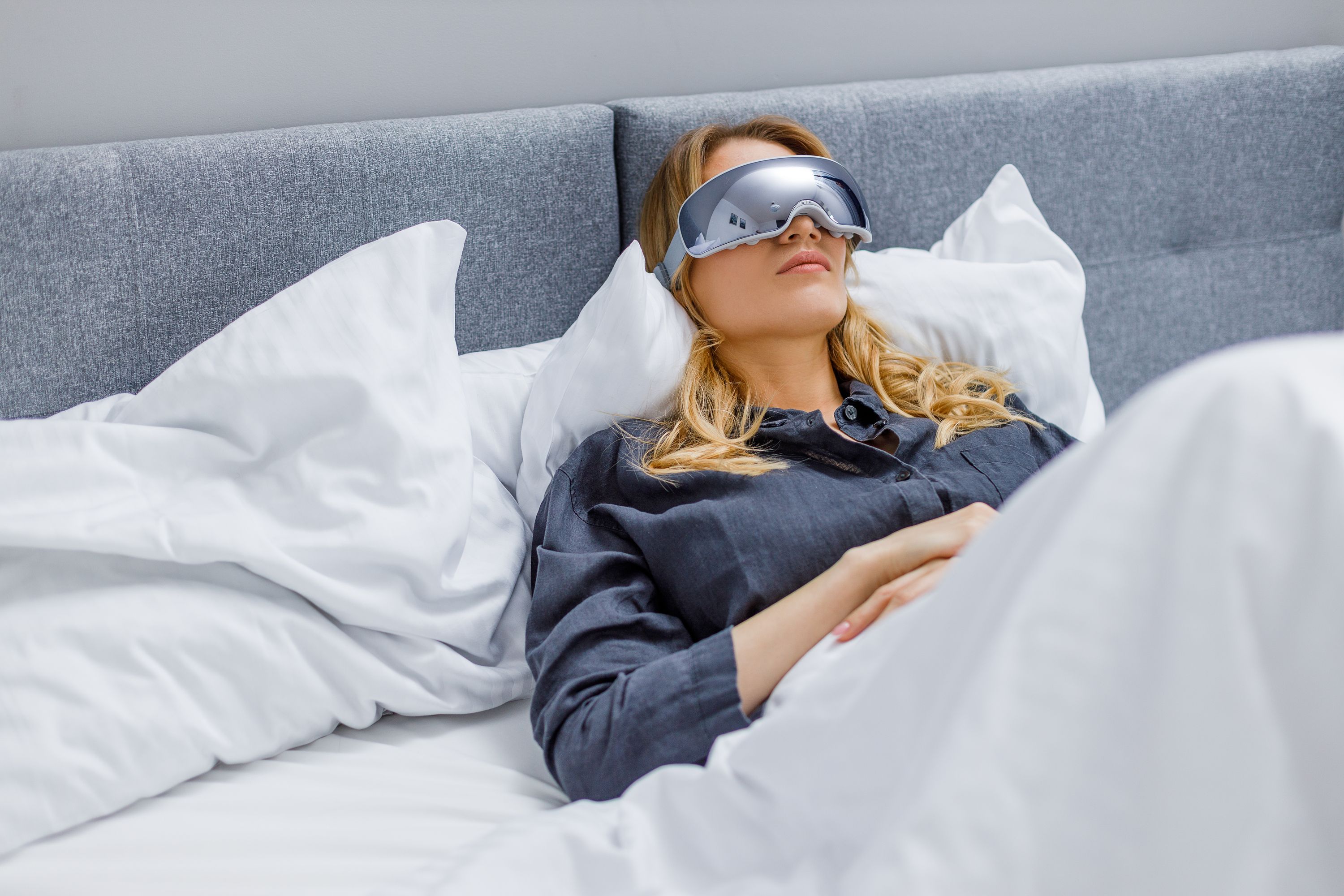 Frau im Bett, Augenmassagegerät auf den Augen. Graues Gerät, graues Band. Auf Kissen und Decke.