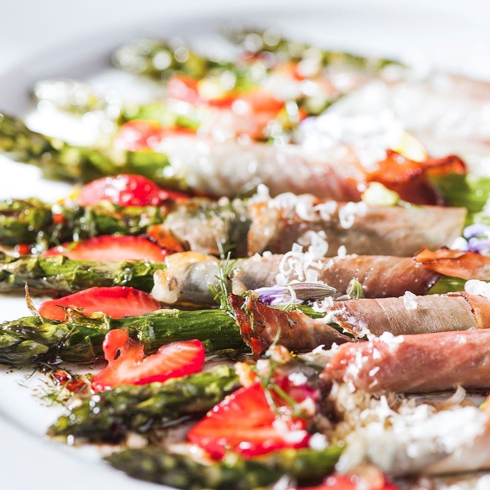Teller mit Spargel, Erdbeeren und Schinkenröllchen. Dekoriert mit Kräutern und Blüten. Auf weißem Hintergrund.