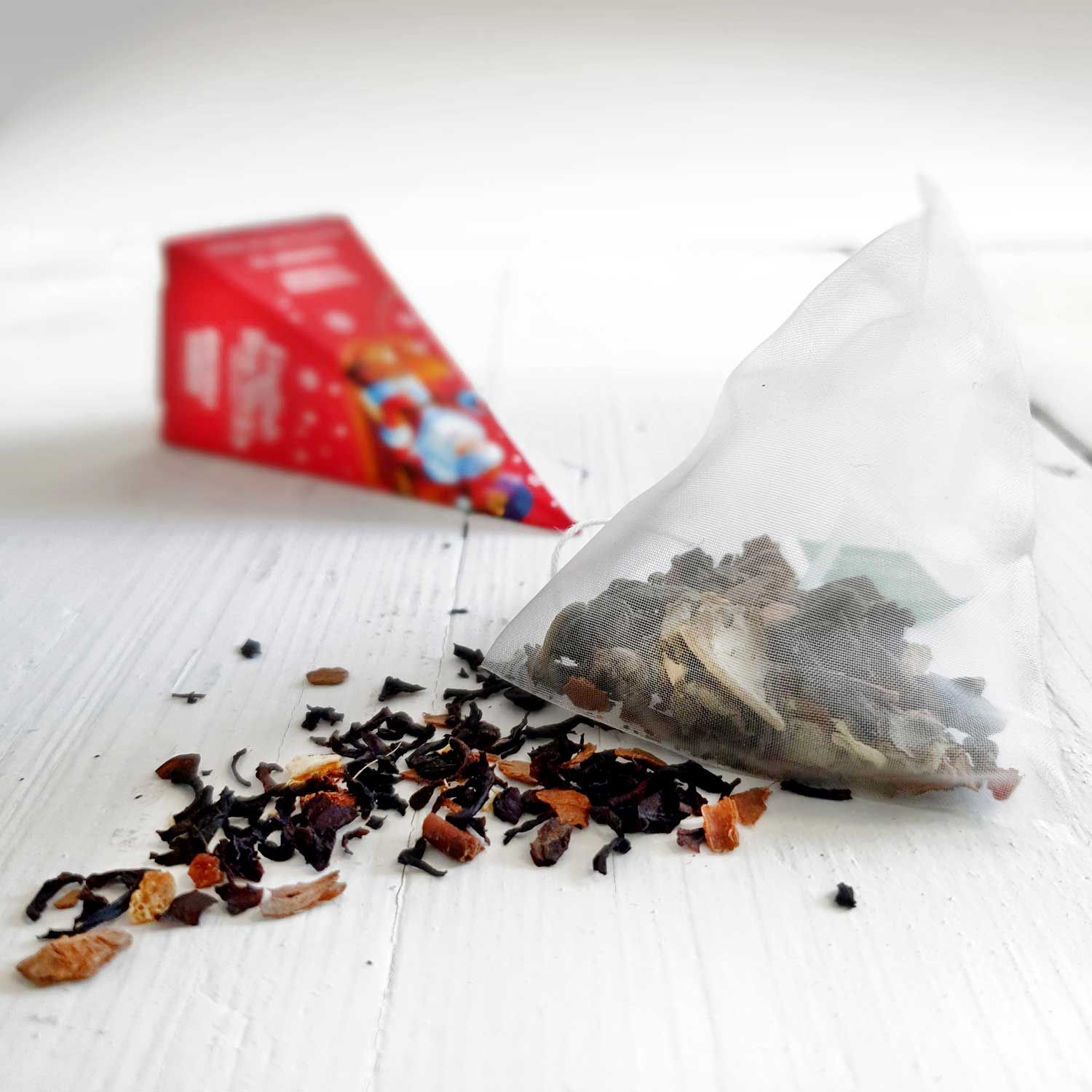 Pyramidenförmiger Teebeutel mit losem Tee. Roter Karton im Hintergrund. Aufschrift nicht lesbar.