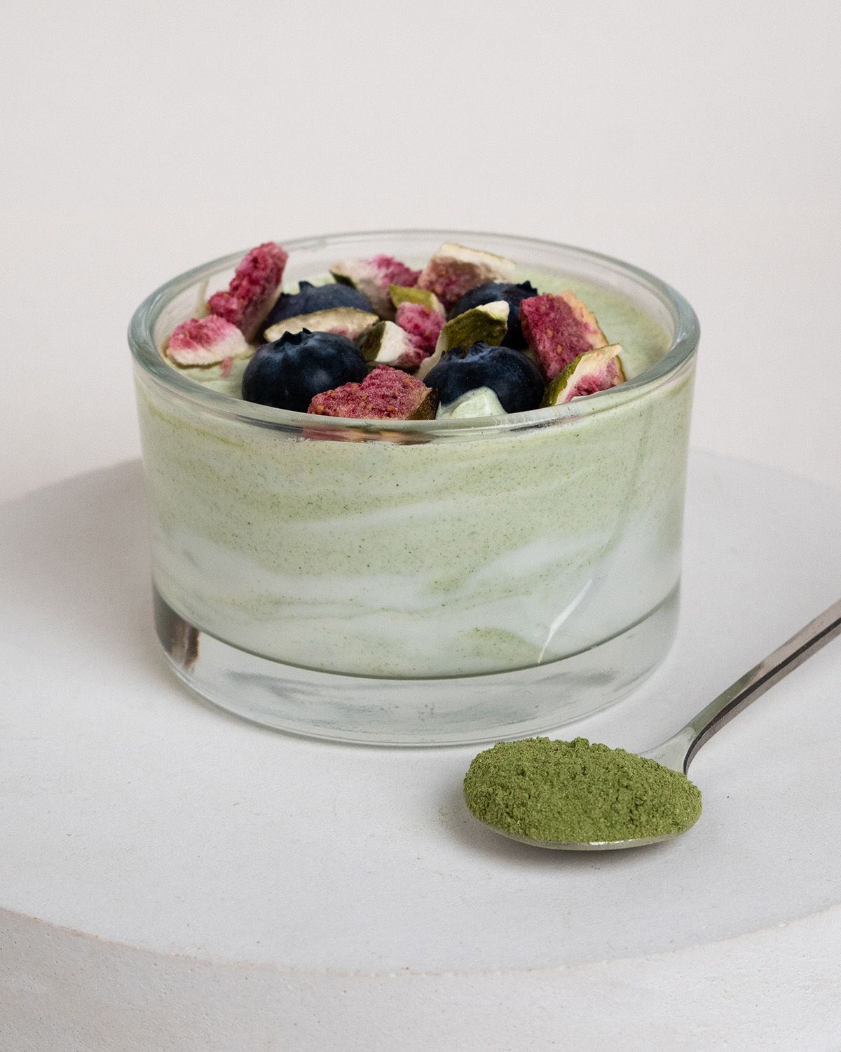 Grünes Weizengraspulver in einem Glas mit Früchten und Beeren garniert, daneben ein Löffel Pulver.