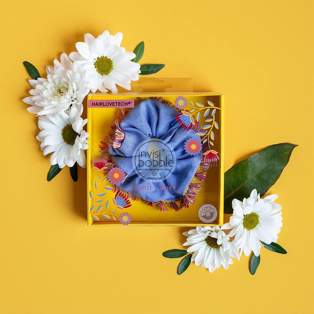 Hola Lola Sprunchie Scrunchie in hellblau mit Fransen in gelber Verpackung, umgeben von Blumen.