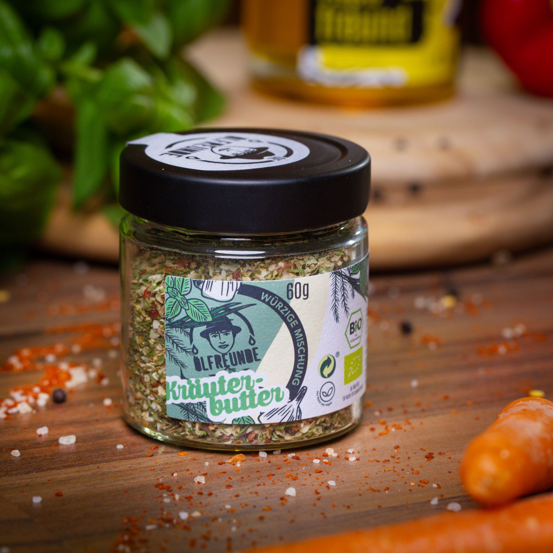 Glas mit Kräuterbuttergewürz auf Holz. Schwarzer Deckel mit Logo. Aufschrift: Kräuterbutter, 60g, Bio-Siegel. Karotte und Gewürze daneben.