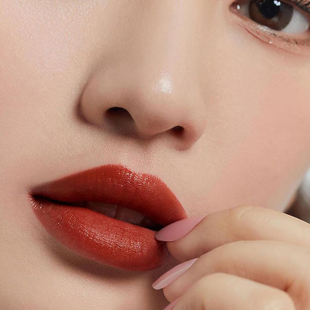 Nahaufnahme von Lippen, die mit einem rotbraunen Lippenstift bemalt sind. Eine Hand mit rosa lackierten Nägeln berührt die Lippen.