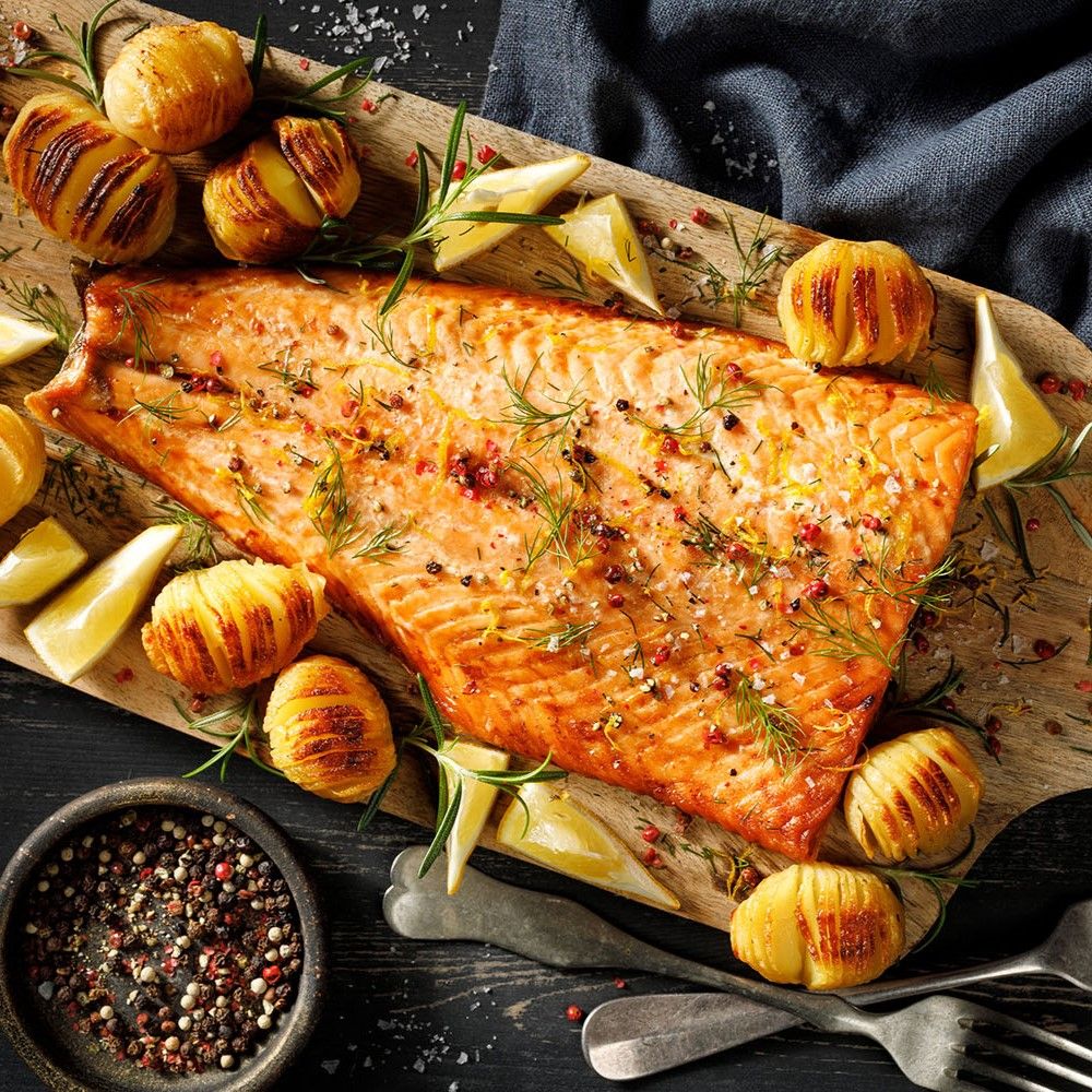 Gebratenes Lachsfilet auf Holzbrett. Dazu Kartoffeln, Zitronen, Kräuter und Pfefferkörner. Auf dunklem Untergrund, mit Besteck.