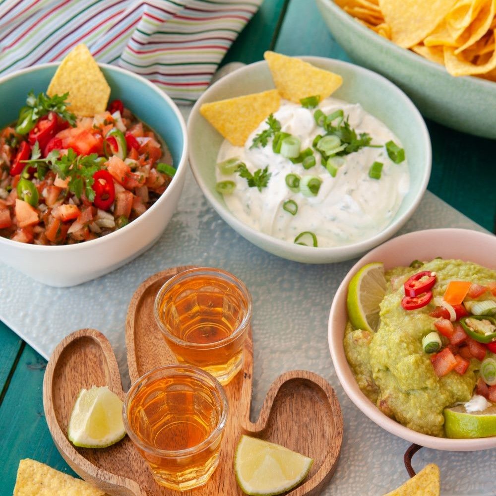 Verschiedene Dips in Schalen, Tortilla-Chips, Limetten und Tequila-Shots auf einem Tisch.