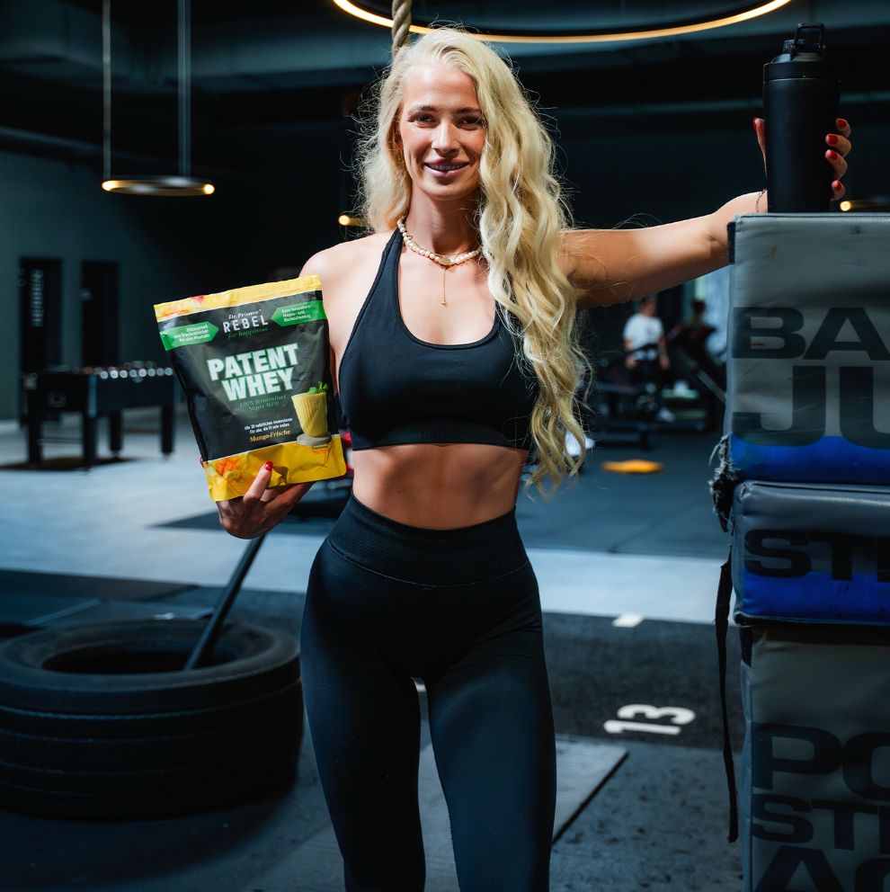 Frau im Fitnessstudio hält eine Tüte mit Produkt. Aufdruck: Rebel for Happiness, Patent Whey. Sie trägt Sportkleidung.