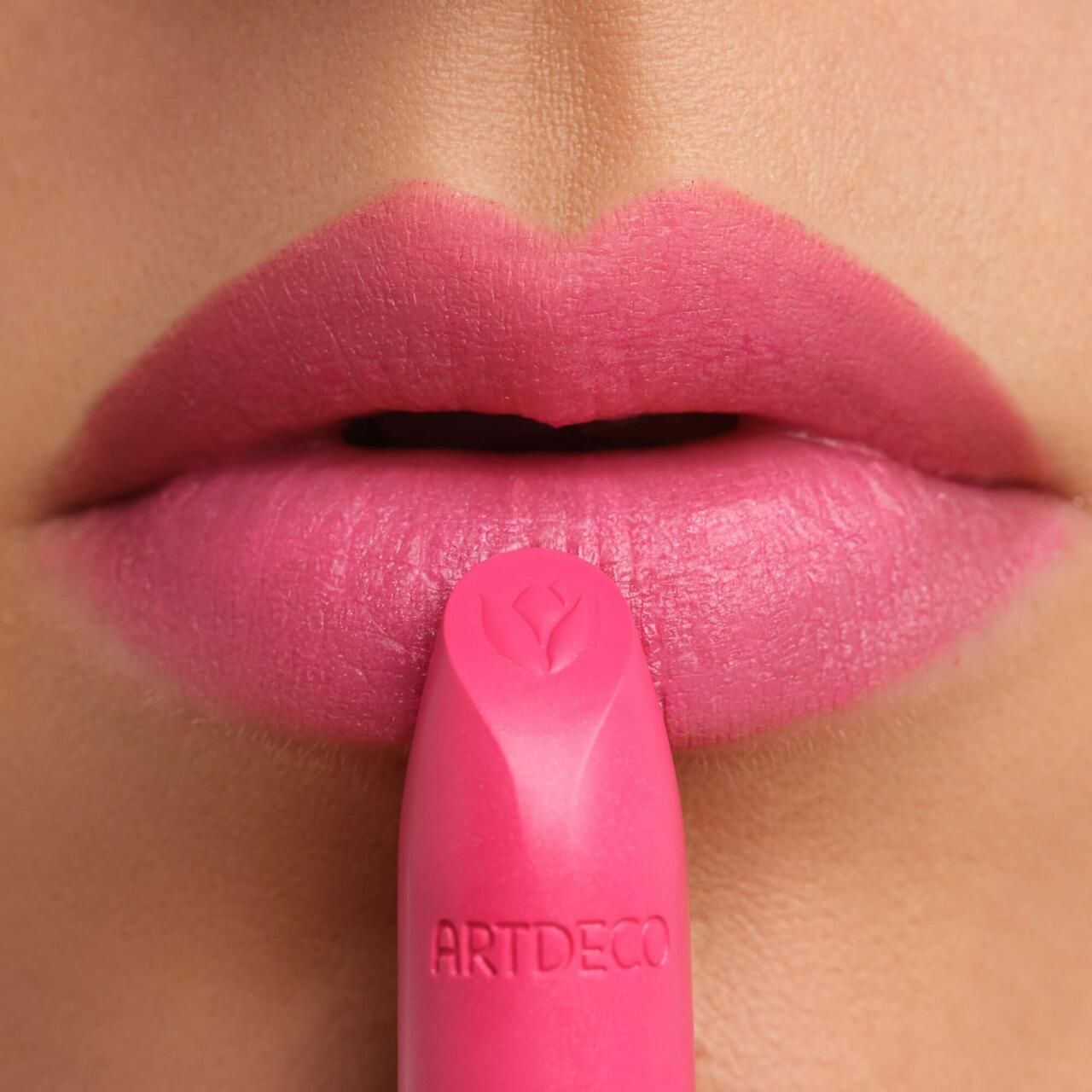 Pinkfarbener Lippenstift vor geöffneten Lippen. Artdeco-Logo auf dem Lippenstift. Nahaufnahme.