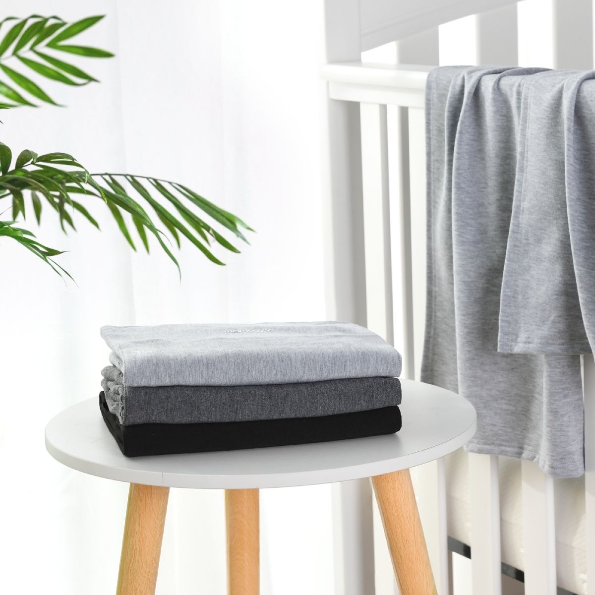 Drie gevouwen draagdoeken in grijstinten en zwart, gestapeld op een kleine witte tafel. Een andere doek hangt over de rugleuning van een witte wieg. Op de achtergrond zijn groene bladeren te zien.
