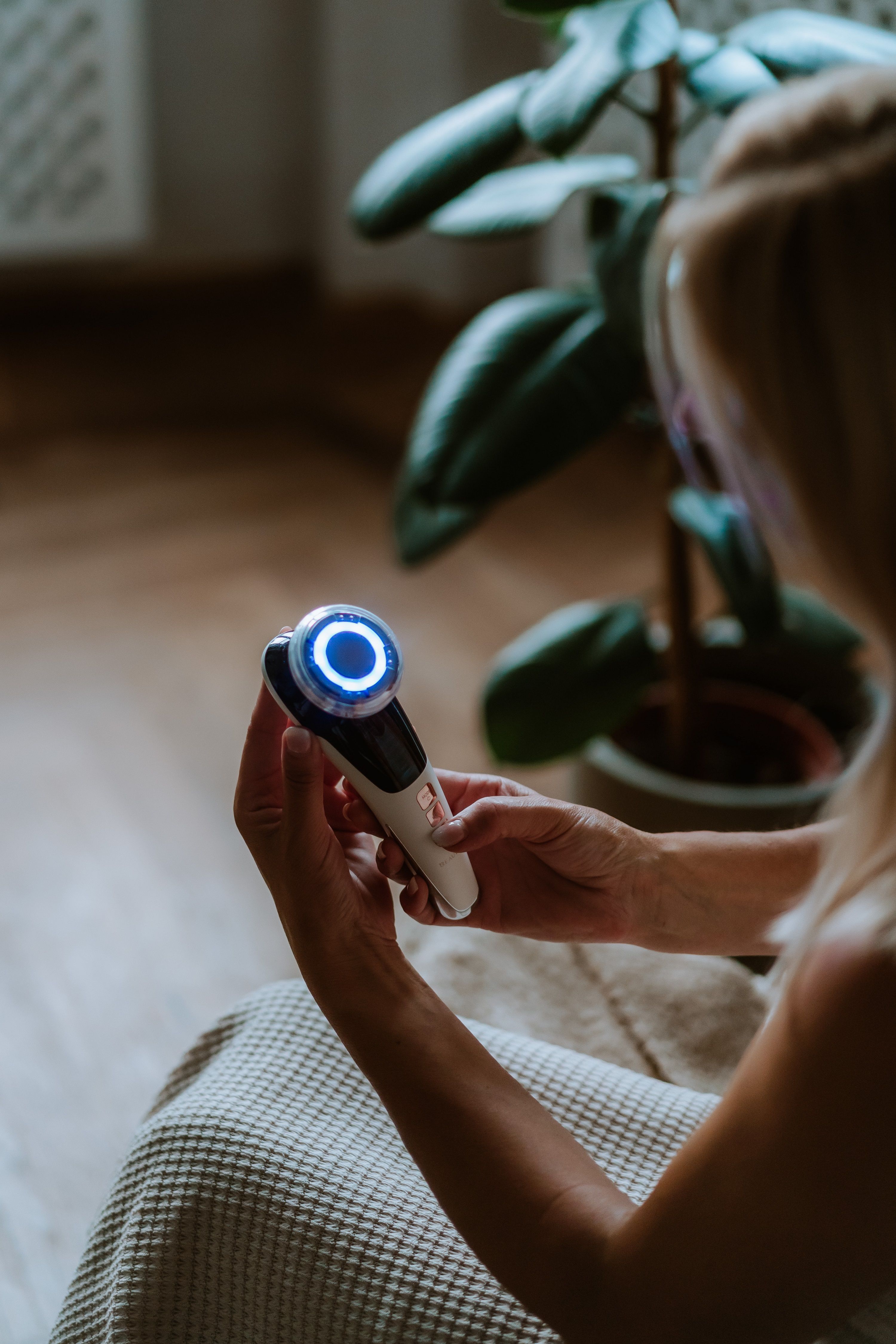 Person hält ein weißes Mesotherapiegerät. Blaue LED-Beleuchtung. Hintergrund: Pflanze und Holzfußboden.