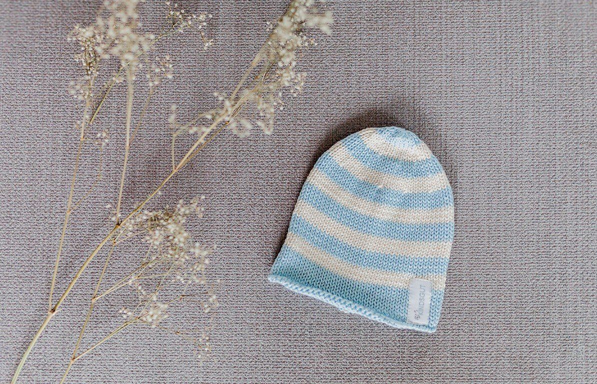 Hellblau-weiß gestreifte Kindermütze auf grauem Untergrund. Kindsgut-Label. Trockenblumen.