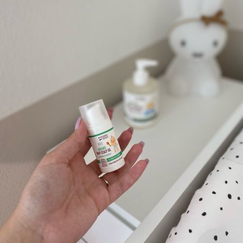 Hand hält eine Flasche Wooden Spoon Bio-Babykopfhautöl. Im Hintergrund: weitere Flasche, Wickeltisch.