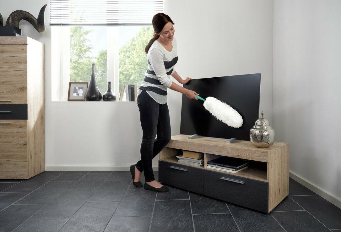Frau reinigt Fernseher mit Staubwedel. Weißer Staubwedelkopf, grüner Griff. Möbel im Hintergrund.