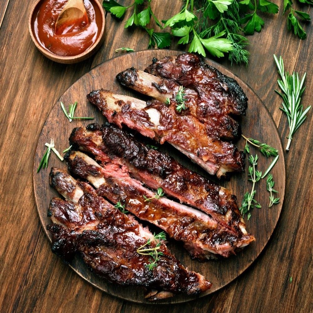 Holzbrett mit gegrillten Rippchen. Rosmarin und Thymian als Garnierung. Schale mit Sauce.