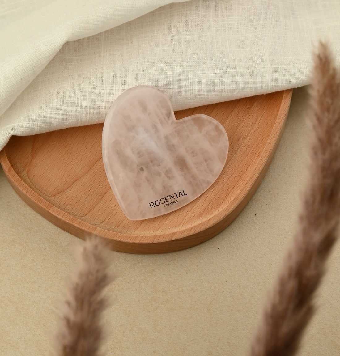 Herzförmiges Gua Sha aus Rosenquarz auf Holztablett. Marke Rosental Organics. Produkt von getrockneten Pflanzen umgeben.