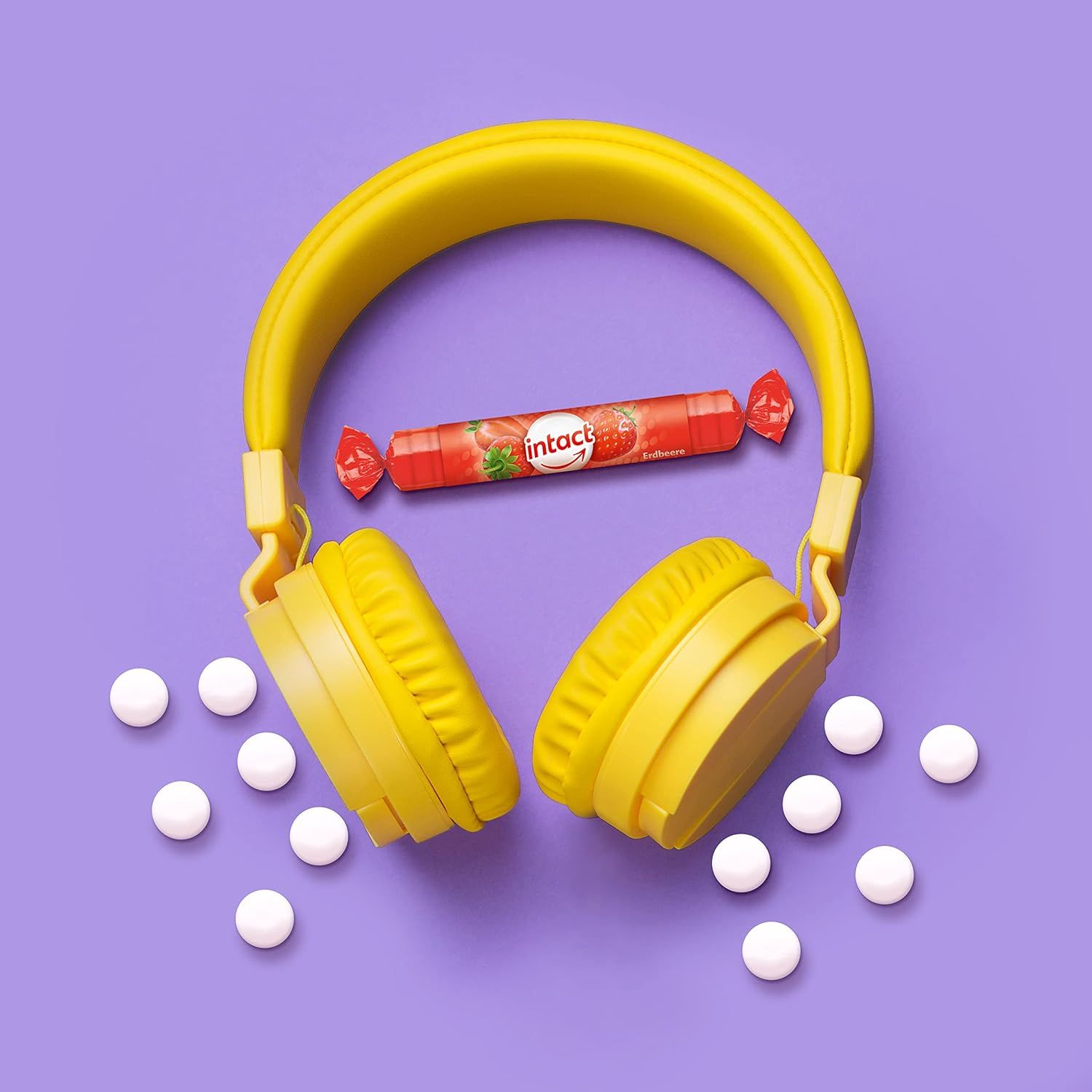 Ein Erdbeer-Traubenzucker-Rolle neben gelben Kopfhörern und weißen Tabletten auf violettem Hintergrund.