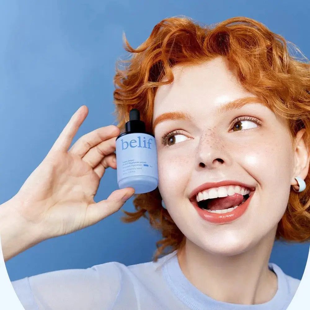 Frau mit roten Haaren hält eine Belif Super Drops Serumflasche vor blauem Hintergrund.