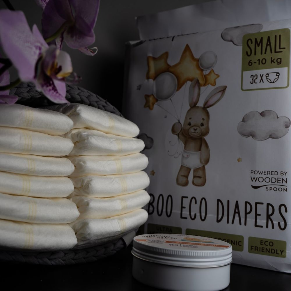 Babywindeln in einem Korb, daneben eine Creme in einer Dose. Im Hintergrund: Packung Babywindeln. Aufdruck: Hase mit Luftballons, Sterne, Wolken. Text: Bamboo Eco Diapers.