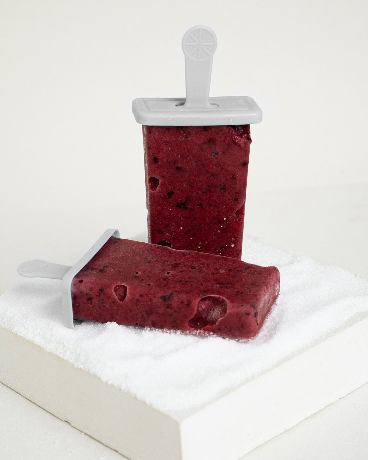 Zwei gefrorene Fruchteis-Popsicles auf einem weißen Handtuch. Einer steht, einer liegt.
